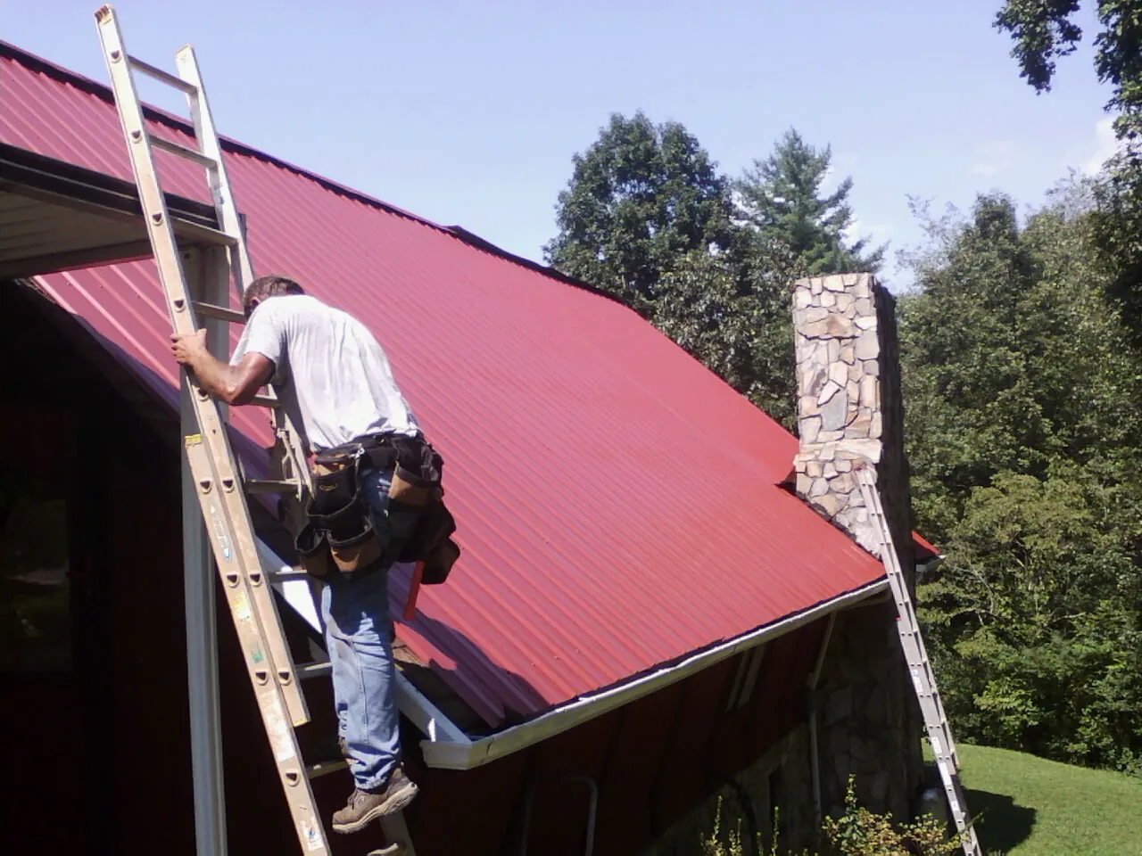 Roofer on a ladder beside a red metal roof and stone chimney — Soffit & Fascia Repair by Roofing Brothers in Ontario